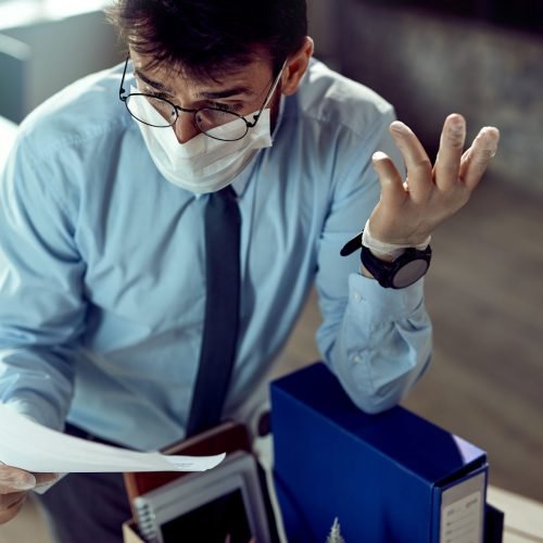 Fired businessman wearing face mask and feeling frustrated while reading a notice in the office.